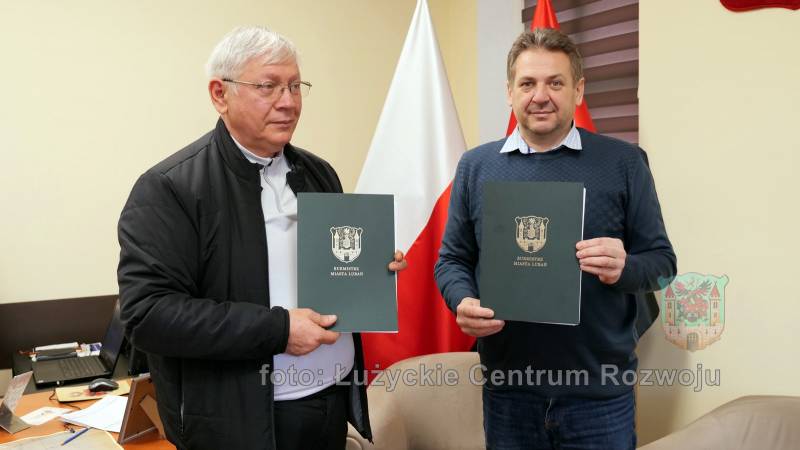 dwóch mężczyzn stoi w pomieszczeniu na tle flag Polski, w rękach trzymają zielone teczki z herbem Lubania